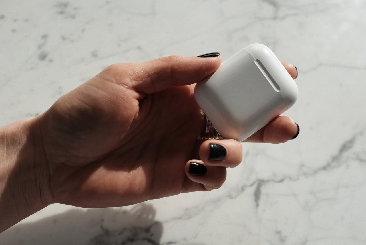 A hand with black nails holding a white wireless earbud case on a marble surface.